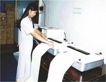 photo: a female RBI staff stands in front of an embosser checking on printed braille materials
