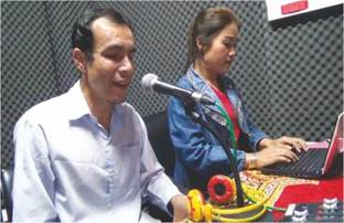 Photo: a visually impaired radio station owner talking through a microphone with a female assistant working on a laptop to his left